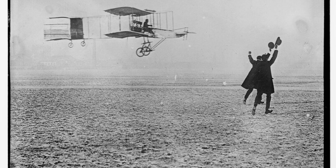 two men wave hats to greet pilot of flying past